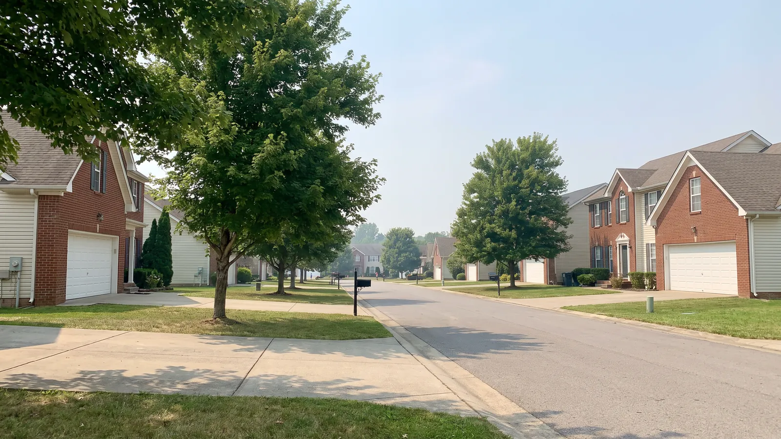 Residential street of 1990s brick and vinyl homes with two-car garages and mature trees in a Clarksville, Tennessee subdivision under a hazy summer sky